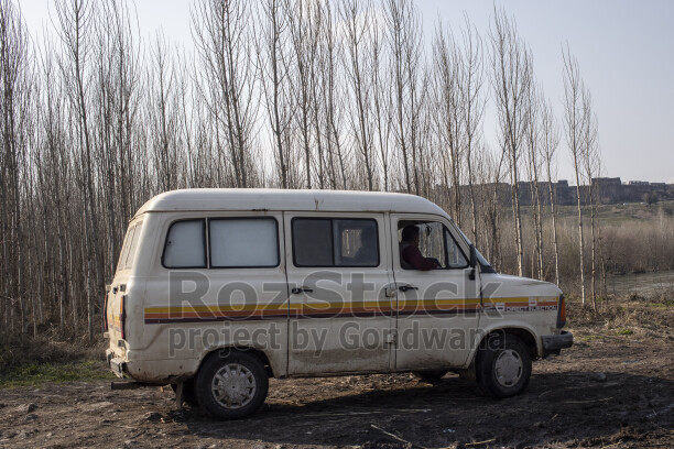Kırsal alanda park halinde eski minibüs, uzun kavak ağaçları ve açık arazi manzarası.