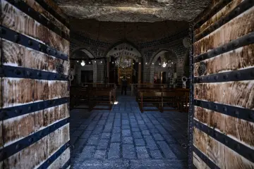 Tarihi kilisenin ahşap kapısından görülen taş kemerli iç mekan, ahşap sıralar ve geleneksel mimari detaylar.