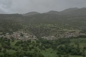 Yeşil vadinin yamaçlarına yayılan yerleşim alanı ve kayalık dağların bulutlu hava altındaki etkileyici manzarası.